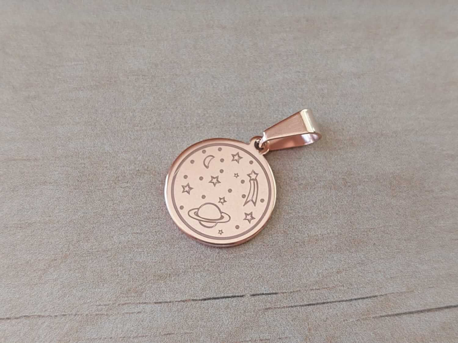 DIJE DE ACERO ROSÉ MEDALLA 18 MM CON SATURNO, LUNA Y ESTRELLAS GRABADAS.
